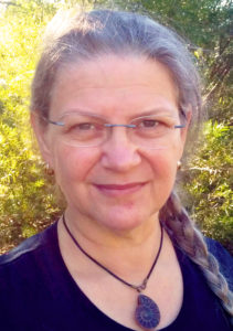 Headshot of a pale-skinned woman outdoors with silvering hair, green leaves in the background. She is wearing spectacles and a neck-choker with a fossilised ammonite pendant.